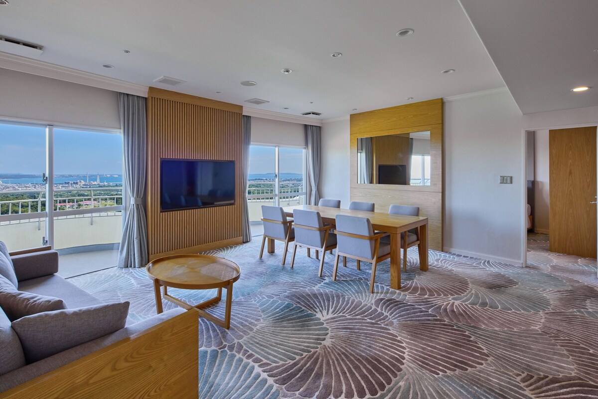 Ein modernes Hotelzimmer mit großem Fenster und Blick auf die Stadt, einem flachbildschirmfernseher an der Wand, einer Essecke mit Tisch und Stühlen sowie einem gemusterten Teppich.
