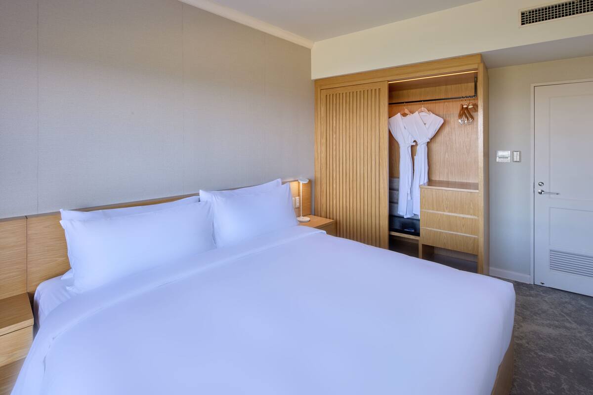 A hotel room with a large bed, wooden headboard, a wardrobe with hanging robes, and a door.