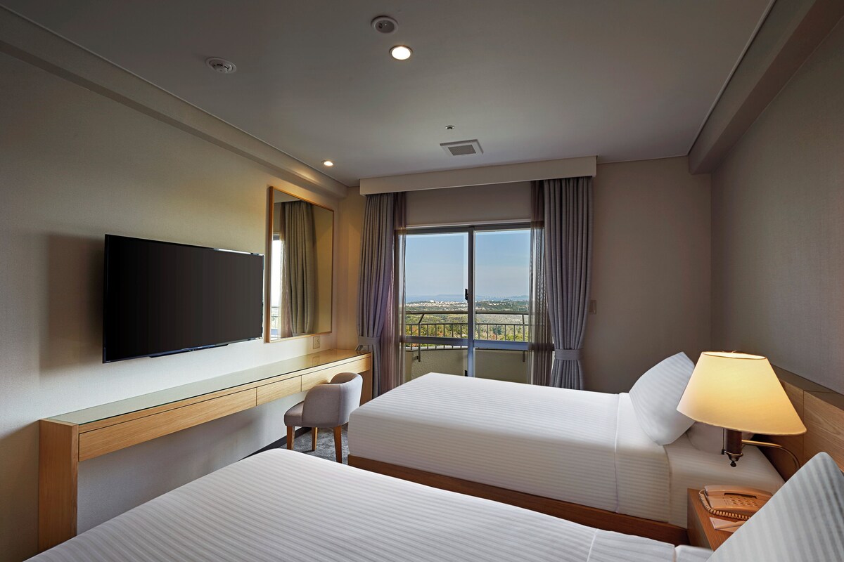 A hotel room with two beds, a desk, a chair, a TV, a window with curtains, and a lamp.