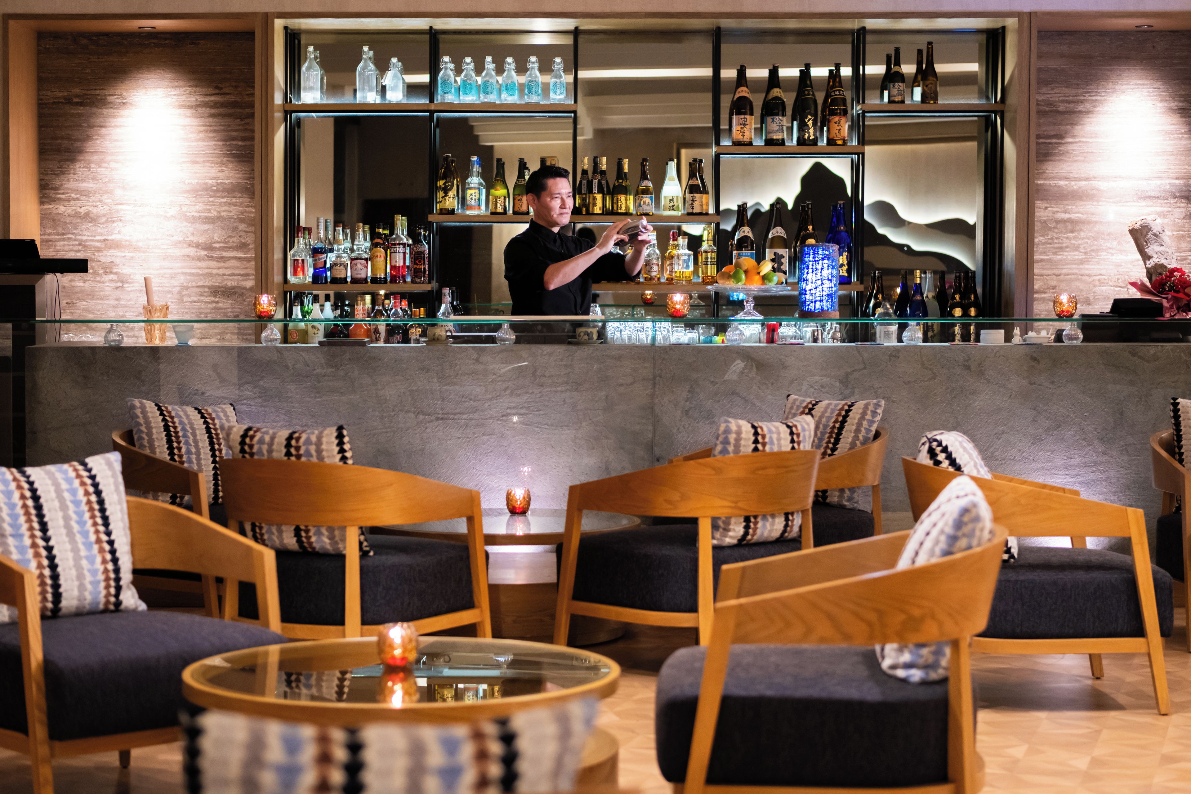 A bar area with a bartender, seating with patterned cushions, and a variety of bottles on shelves.