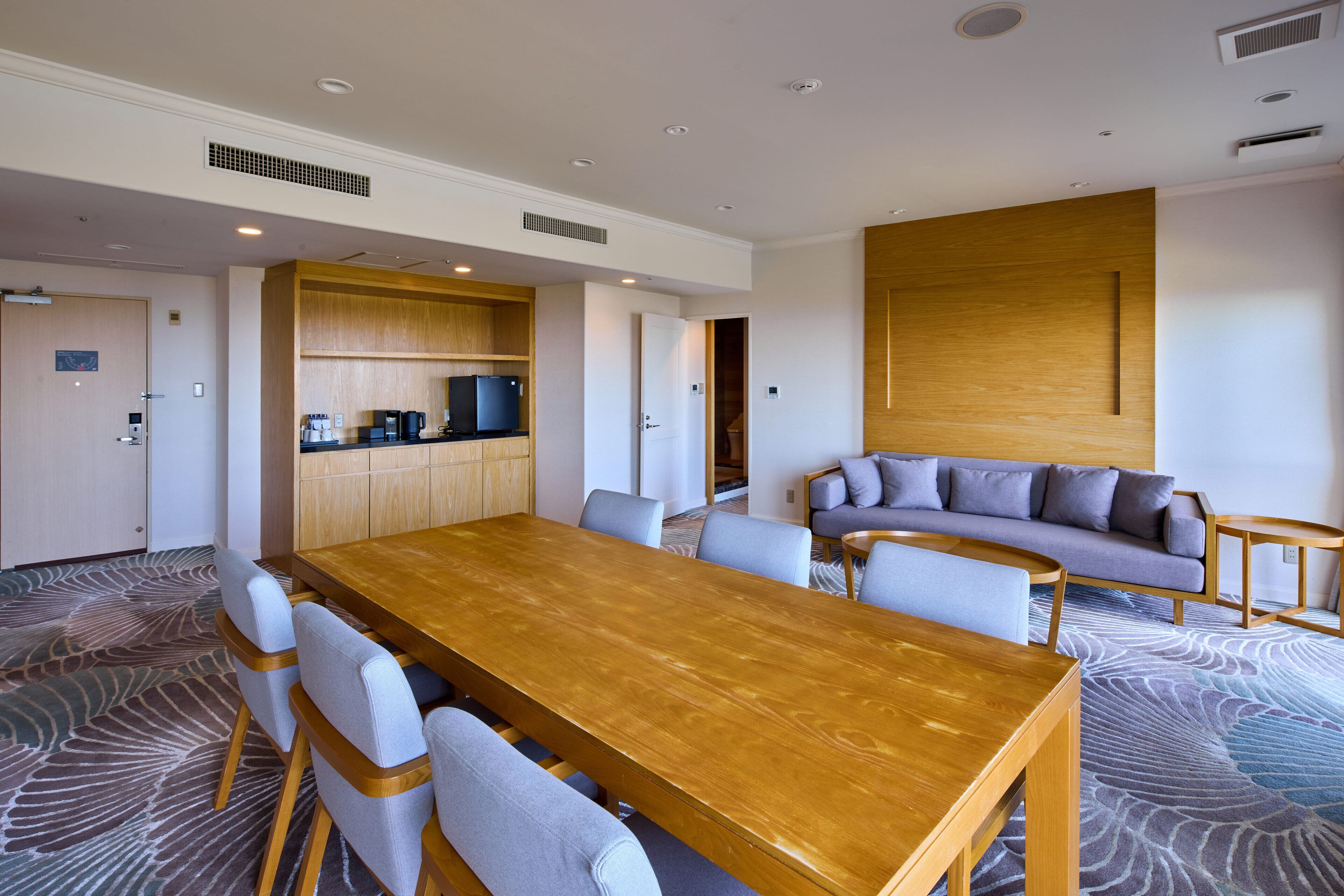 A modern hotel room with a wooden dining table, gray upholstered chairs, a gray sofa, and a patterned carpet.