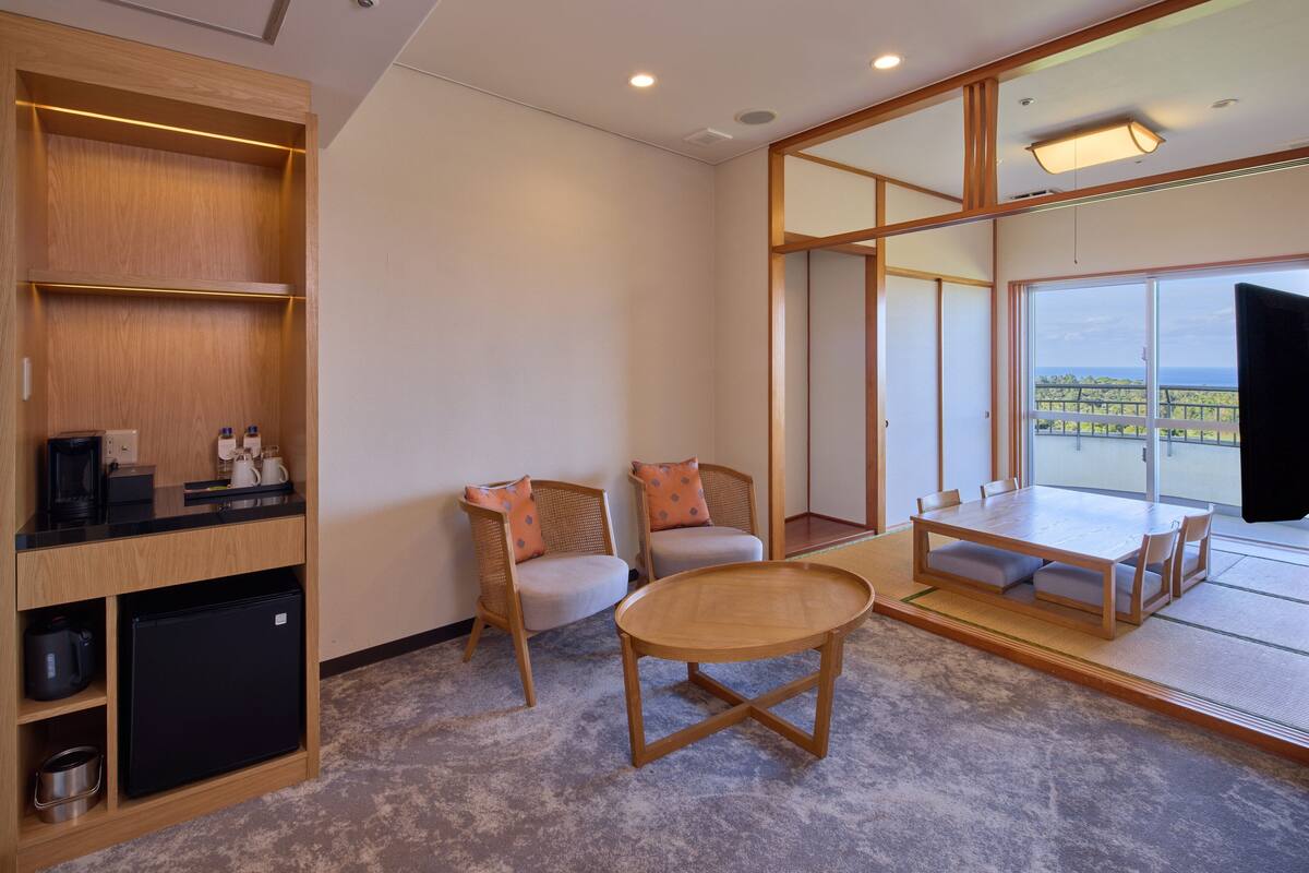 A modern room with a wooden cabinet, a small refrigerator, two chairs, a round table, and a large window offering a view.
