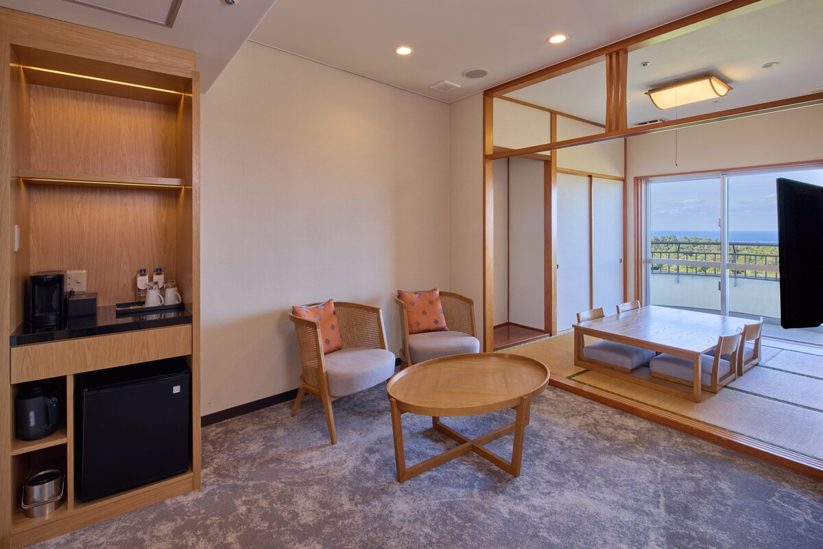 A modern room with a wooden cabinet, a small refrigerator, two chairs, a round table, and a large window offering a view.
