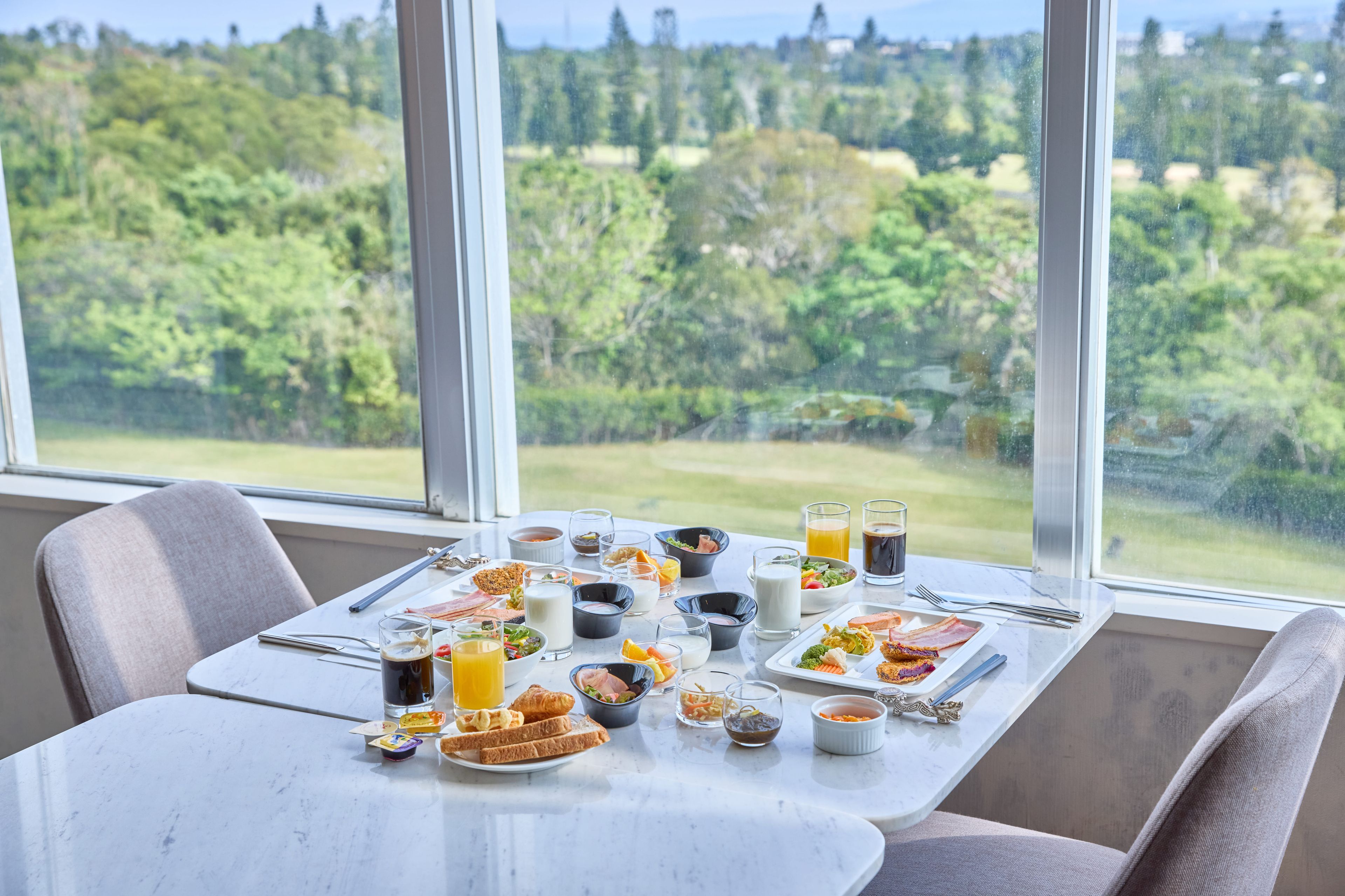 Mesa de café da manhã com diversos pratos e bebidas, em um quarto com grandes janelas com vista para uma paisagem arborizada.