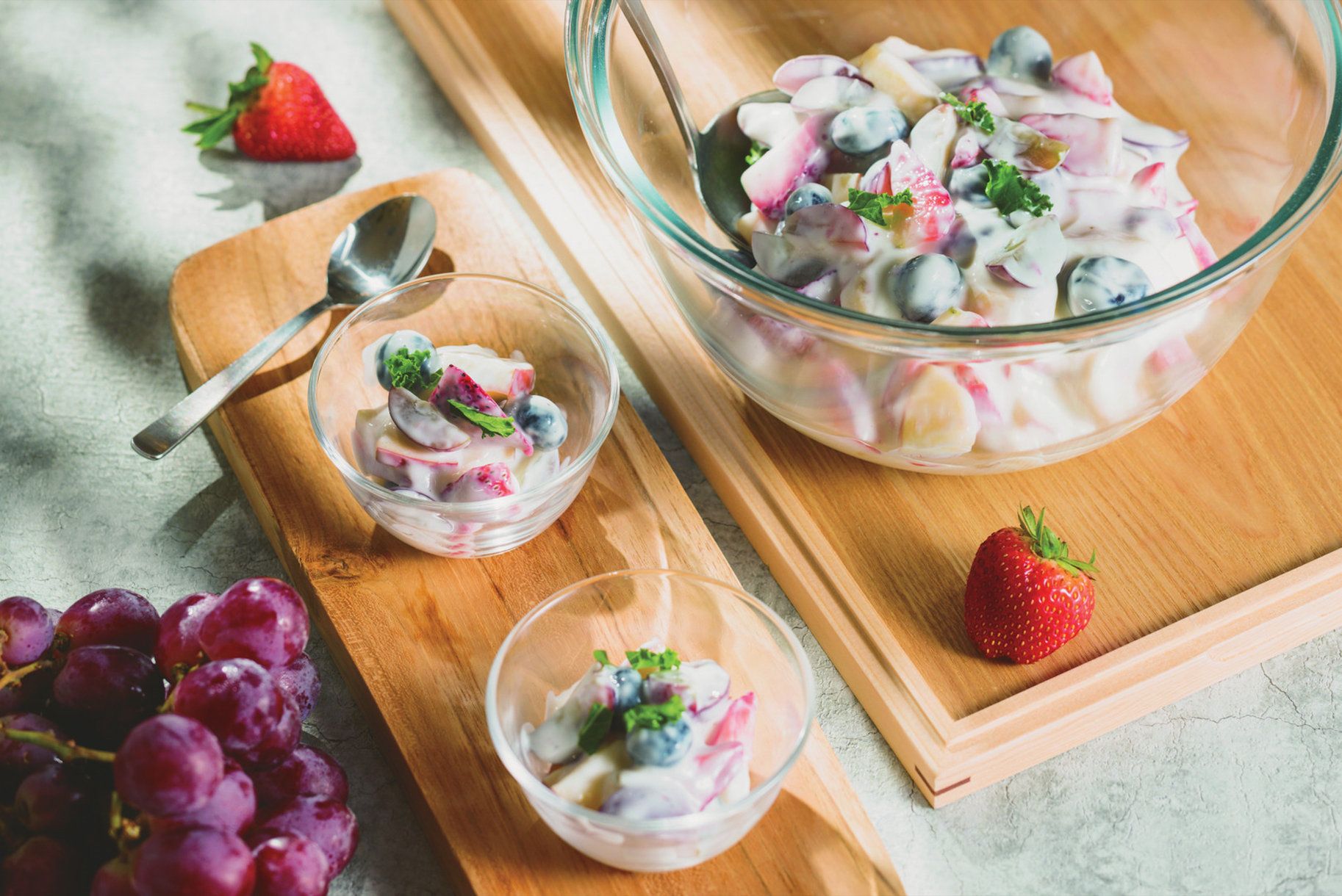 A glass bowl of fruit salad with a spoon on a wooden board.