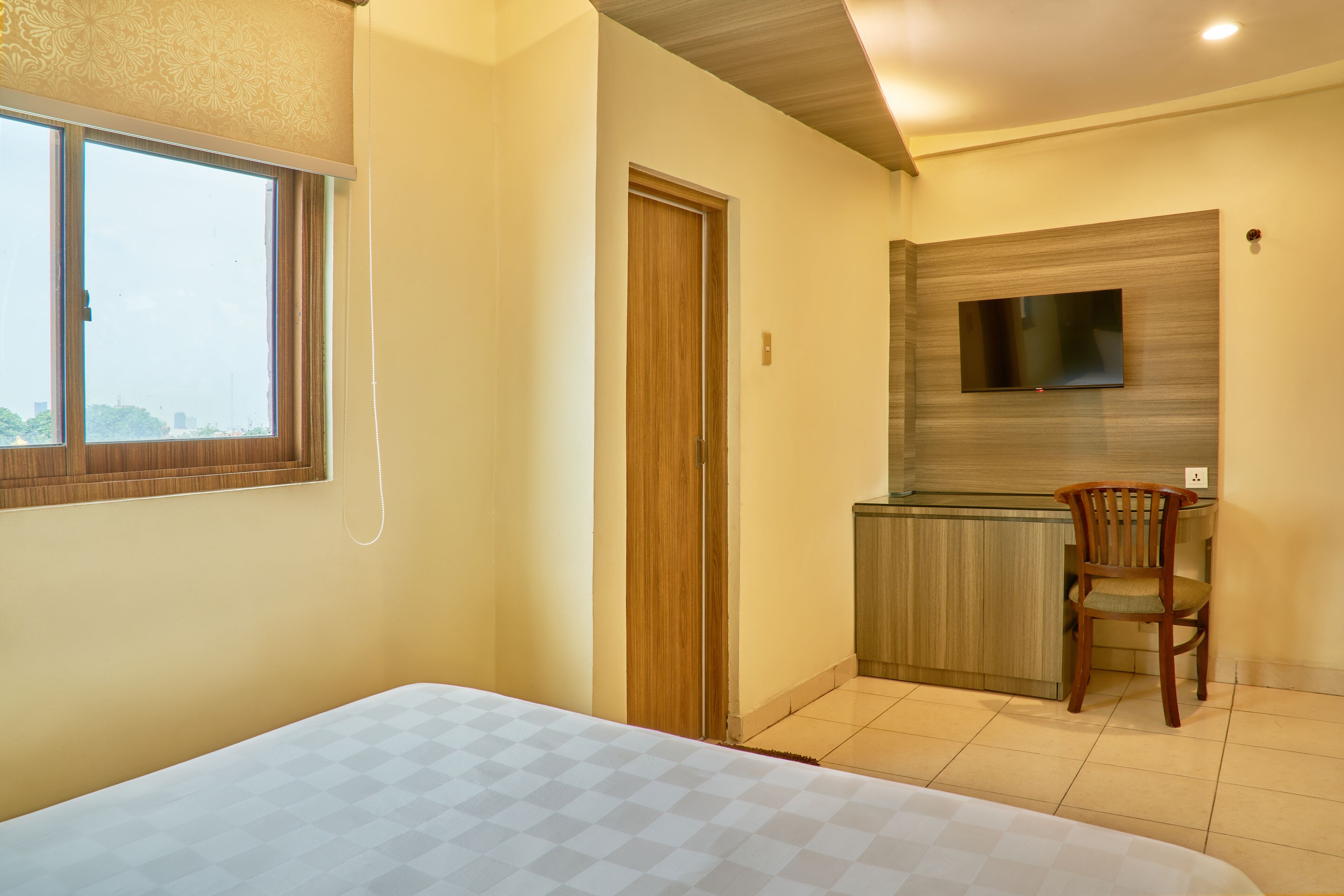 A hotel room with a bed, a small kitchenette, a TV mounted on the wall, and a window with curtains.