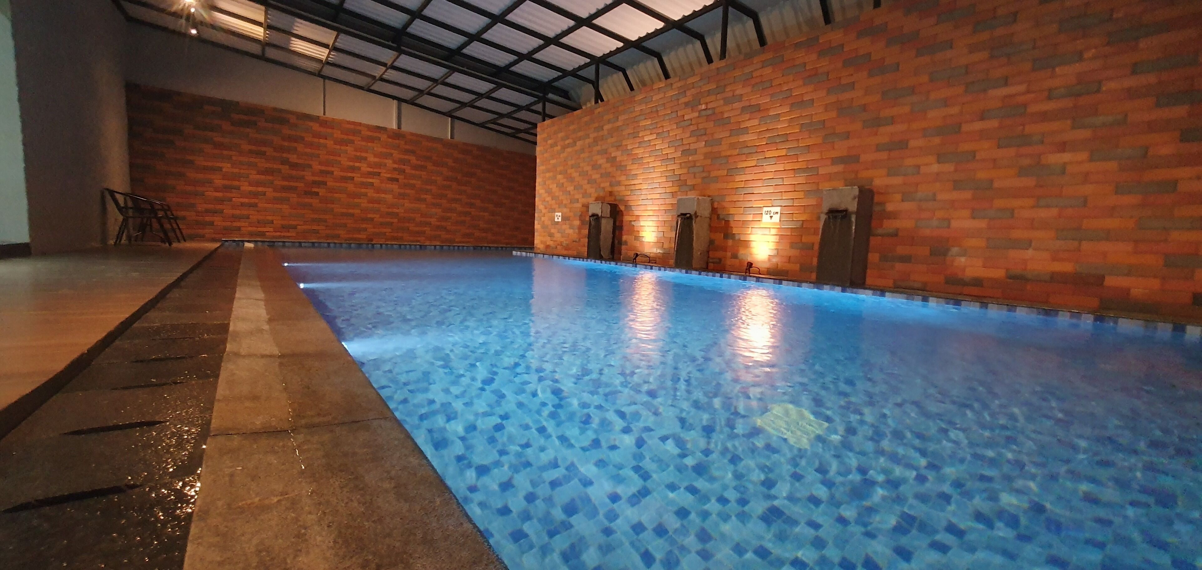Une piscine intérieure dotée d’un sol en mosaïque bleue, entourée de murs en briques et équipée d’un système d’éclairage au plafond.