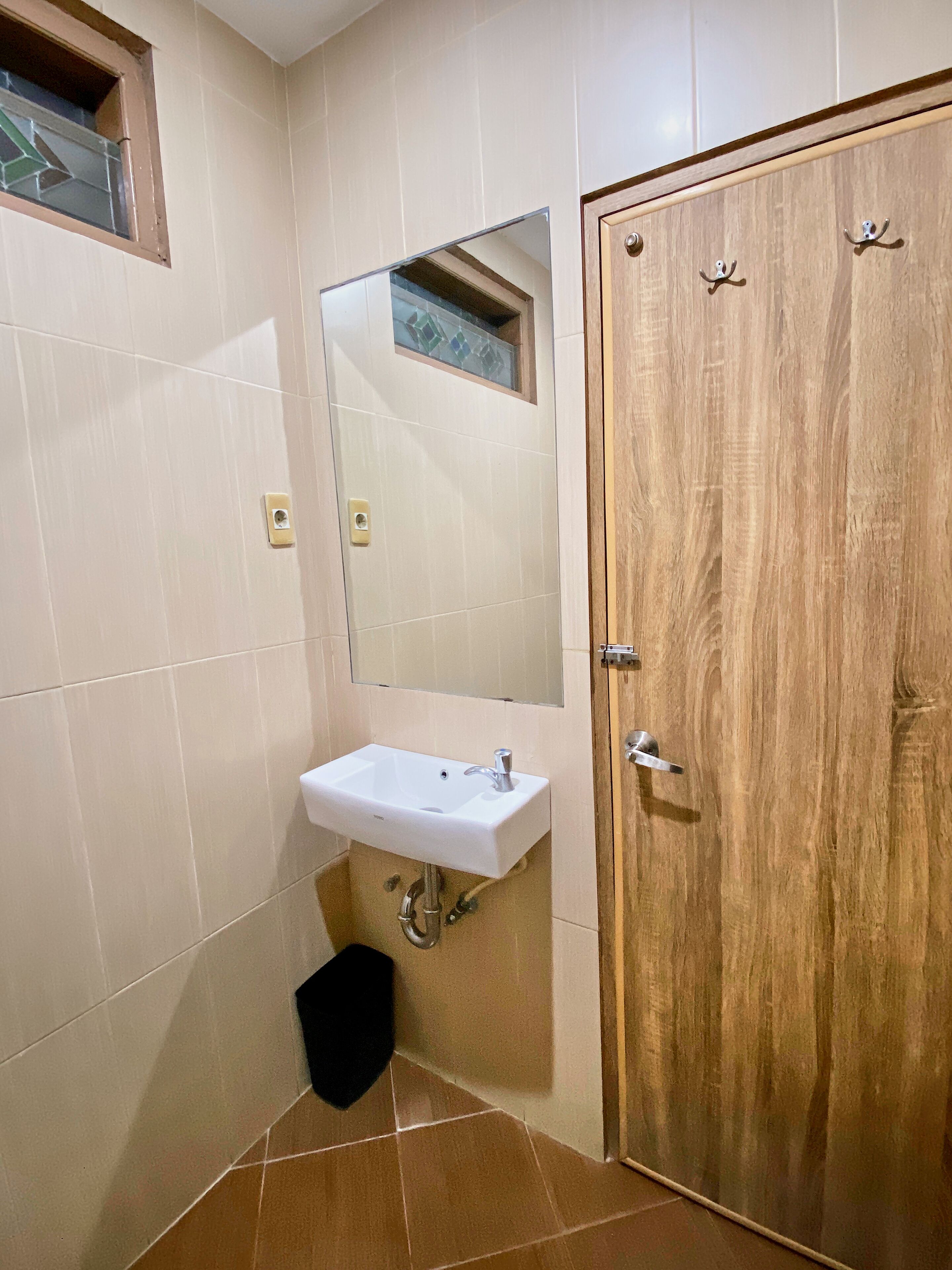 Un baño con puerta de madera, un lavamanos blanco con un espejo encima y un cubo de basura negro.