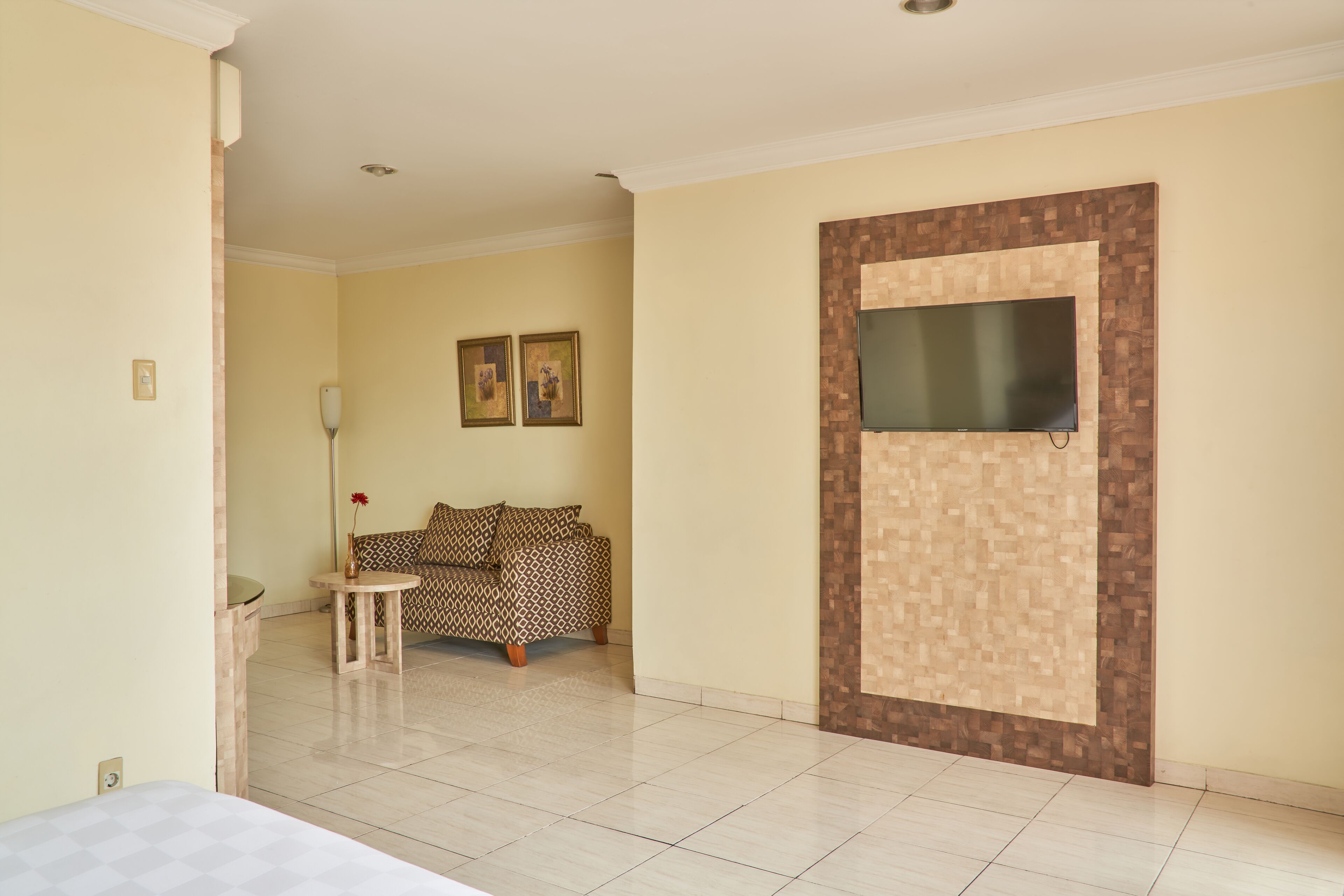 A hotel room with a bed, a small table with a vase, a patterned sofa, and a television mounted on a wall with a wooden design.