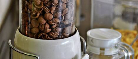 A coffee grinder with a transparent container filled with coffee beans, a small white container with a lid, and a jar with a lid in the background.