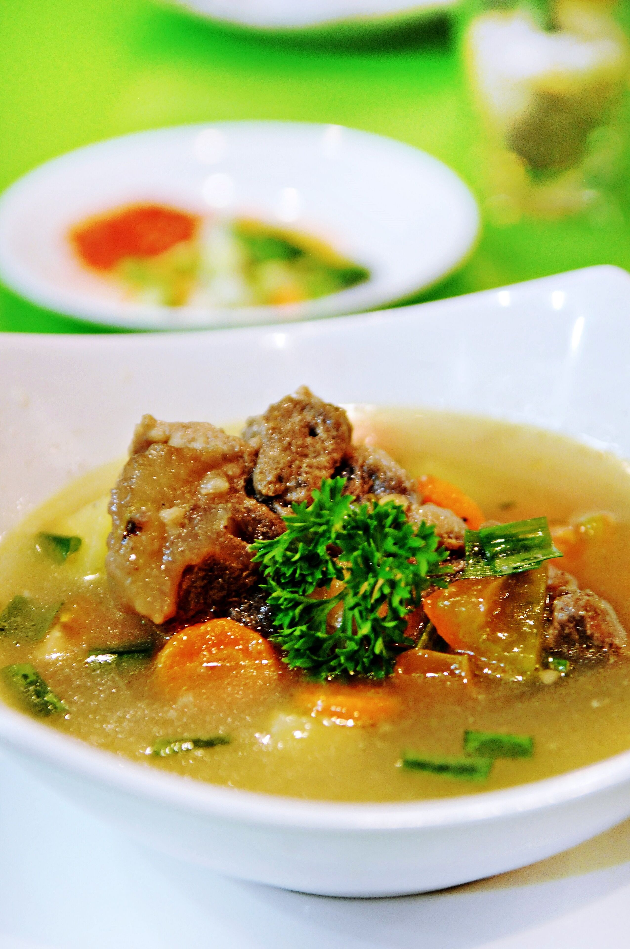 A bowl of soup with meat and vegetables, garnished with parsley, on a green table.