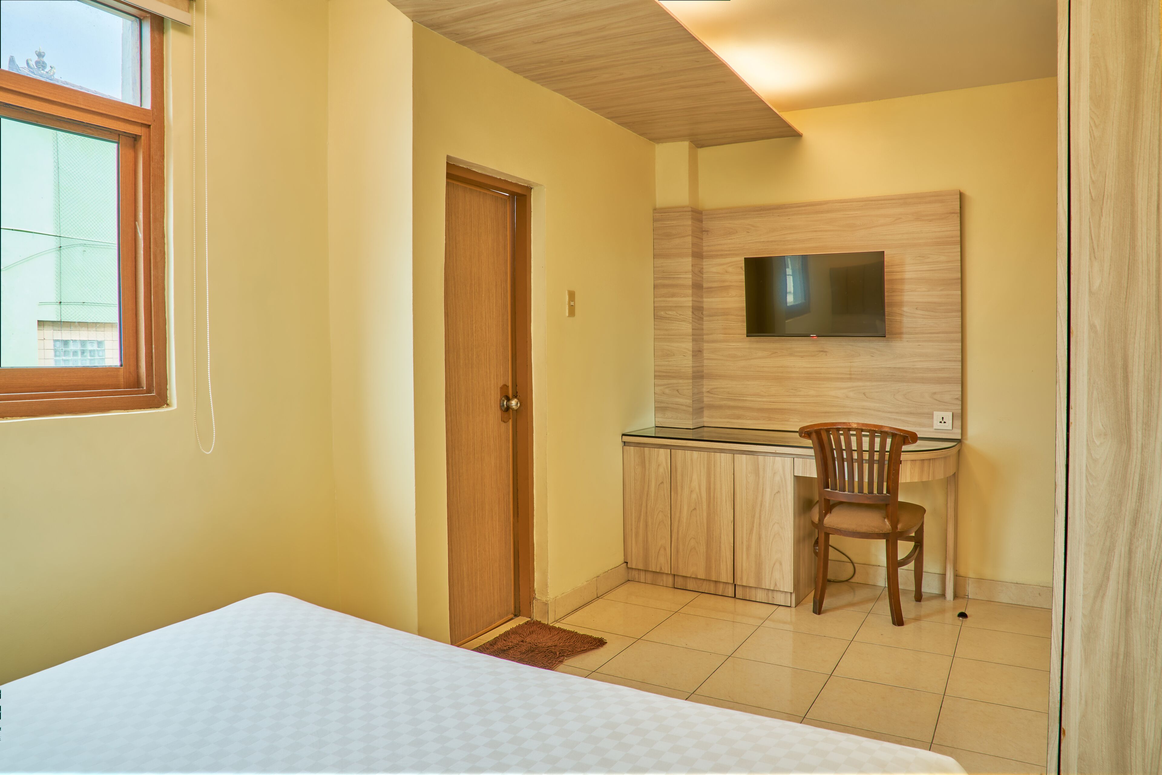A bedroom with a bed, a wooden door, a small rug, a desk with a chair, and a TV mounted on the wall.