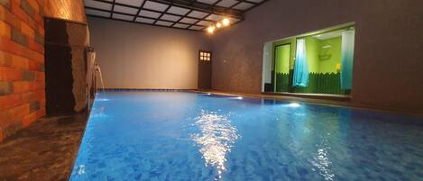 An indoor swimming pool with clear water, a brick wall on one side, and a door leading to a room with a green and white color scheme.