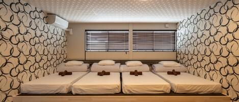 Een hotelkamer met vier enkele bedden, een groot raam met zonwering, een behang met patroon en een hanglamp.