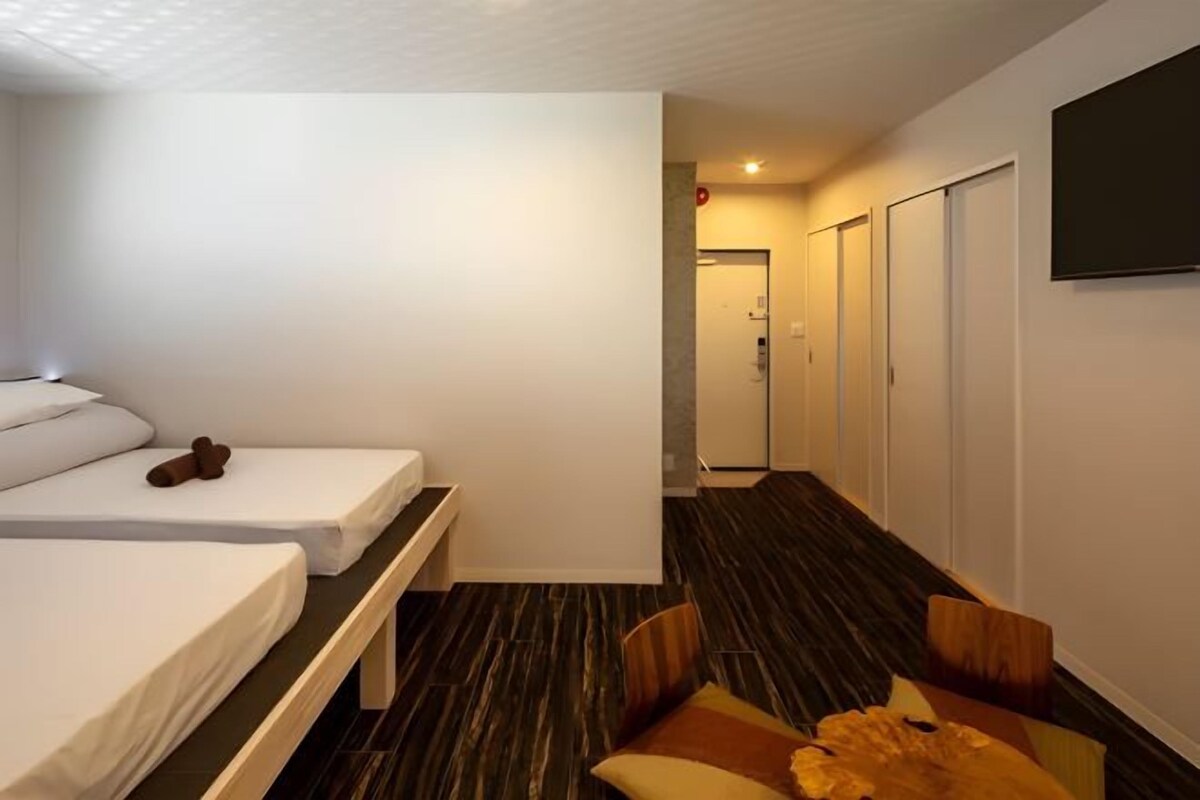 A hotel room with two beds, a bench, a TV, and a hallway leading to a door.