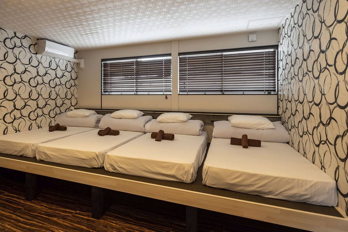 A hotel room with four beds arranged in a row, each with white linens and brown towels. The walls have a geometric pattern, and there are two windows with blinds.