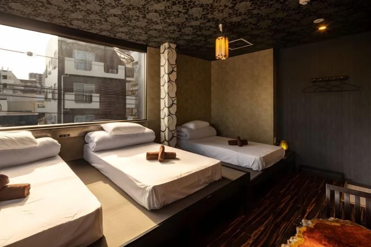 A hotel room with three single beds, a patterned ceiling, a hanging light fixture, and a view of buildings outside.