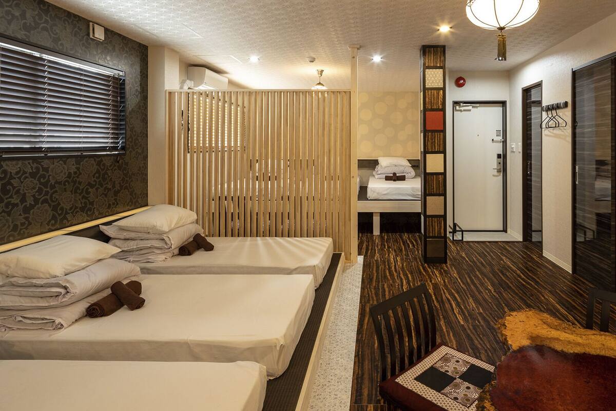 A hotel room with two beds, a wooden partition, a small table with a patterned table runner, and a television on the wall.