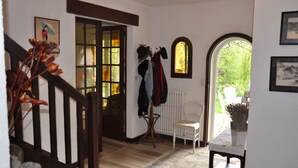 Ein Flur mit einer Holztreppe, einem weißen Stuhl, einem Heizkörper und einem Fenster mit Blick ins Grüne.