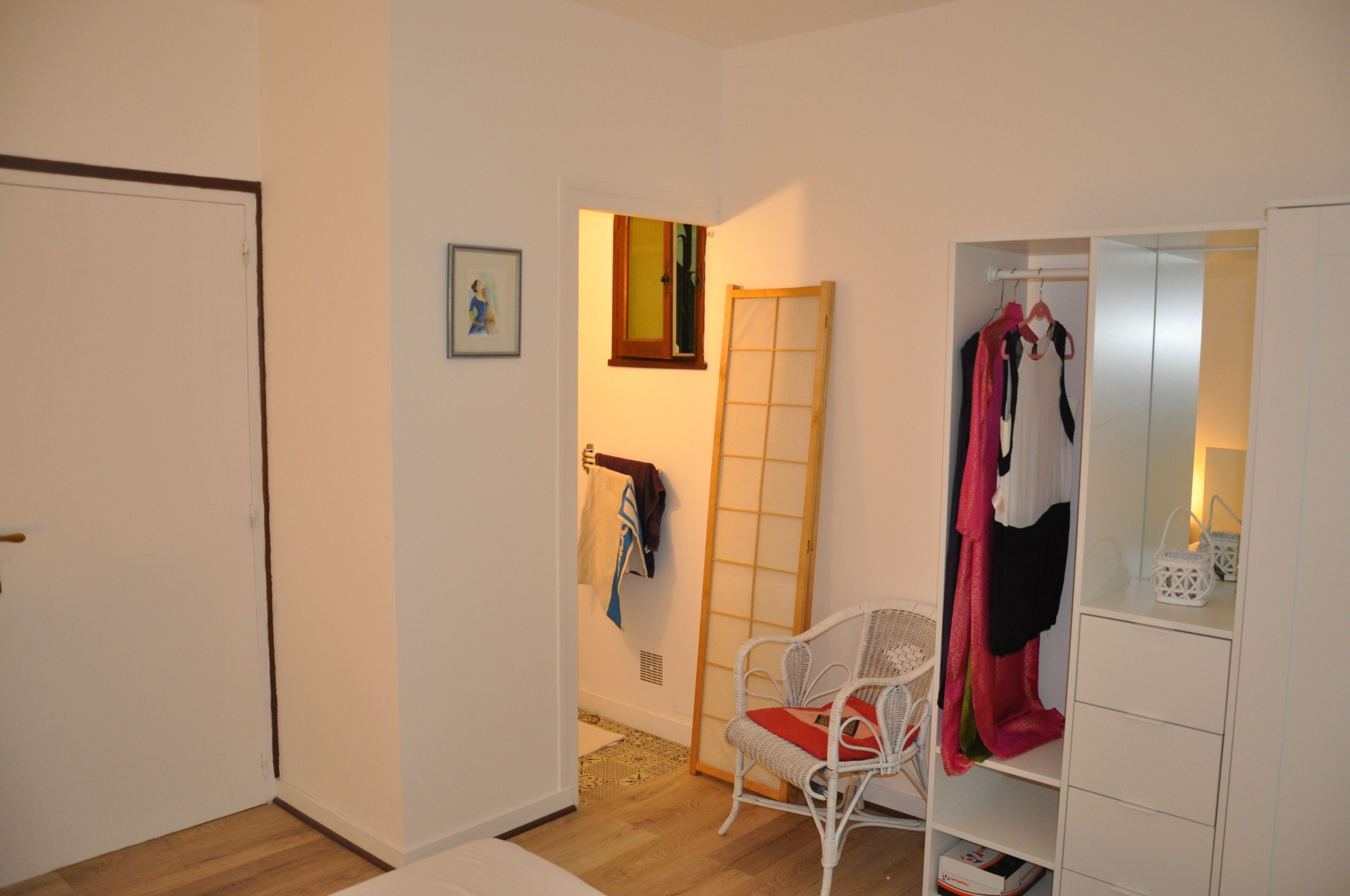 Ein Zimmer mit weißer Tür, einem weißen Stuhl mit rotem Kissen, einem weißen Kleiderschrank und einem kleinen Fenster mit einem Bild an der Wand.