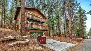 A two-story log cabin with a green roof and a balcony, surrounded by tall pine trees.