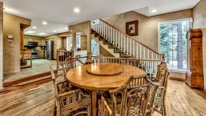 A spacious dining area with a wooden table and wicker chairs, a kitchen in the background, and a staircase leading to another level.