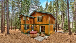 A two-story wooden cabin with a deck and a hot tub, surrounded by a forest.
