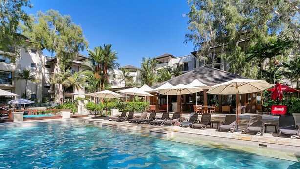 A swimming pool with lounge chairs and umbrellas, surrounded by buildings and palm trees.