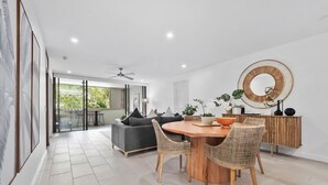 A modern living room with a dining area, featuring a round wooden table, wicker chairs, a grey sofa, and a large mirror on the wall.