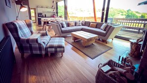 A spacious living area with a large sectional sofa, a coffee table, and a fireplace. There is a dining area with a table and chairs, and a kitchen in the background. The room has large windows and a balcony with outdoor furniture. - A yorkshire dale that sleeps 6 guests  in 3 bedrooms (Richmond)