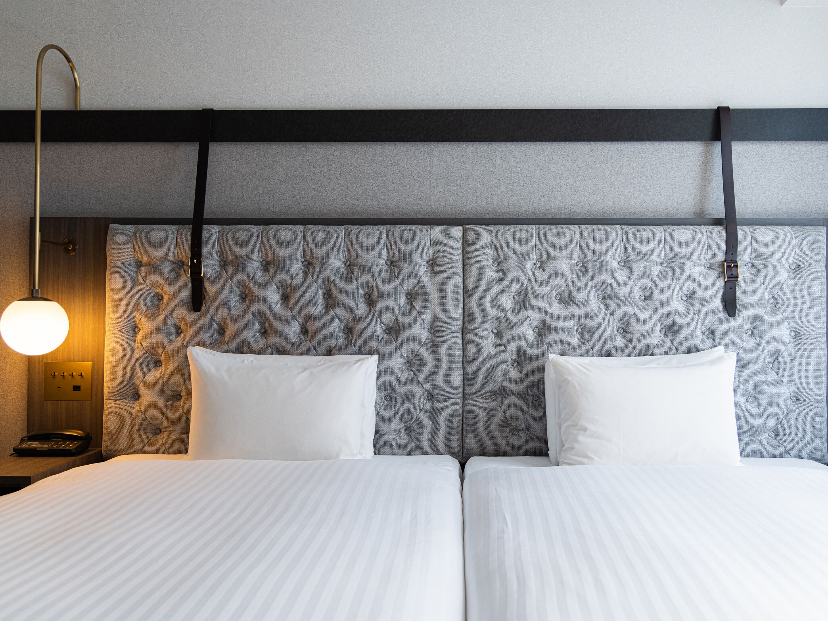 Una habitación de hotel con cabecera capitonada, dos camas con ropa blanca, una lámpara de noche y un teléfono en la mesita.