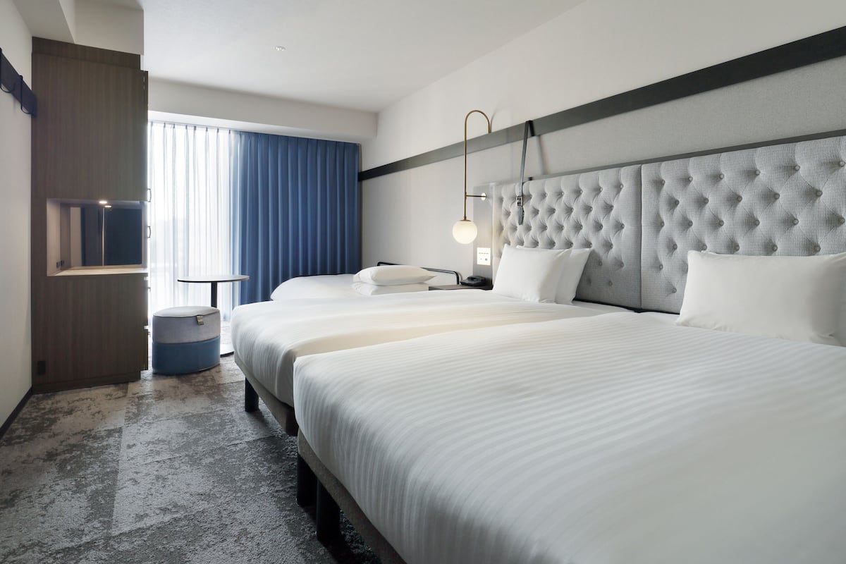 Une chambre d’hôtel avec deux lits, une grande fenêtre avec des rideaux, une table de chevet et une lampe fixée au mur.