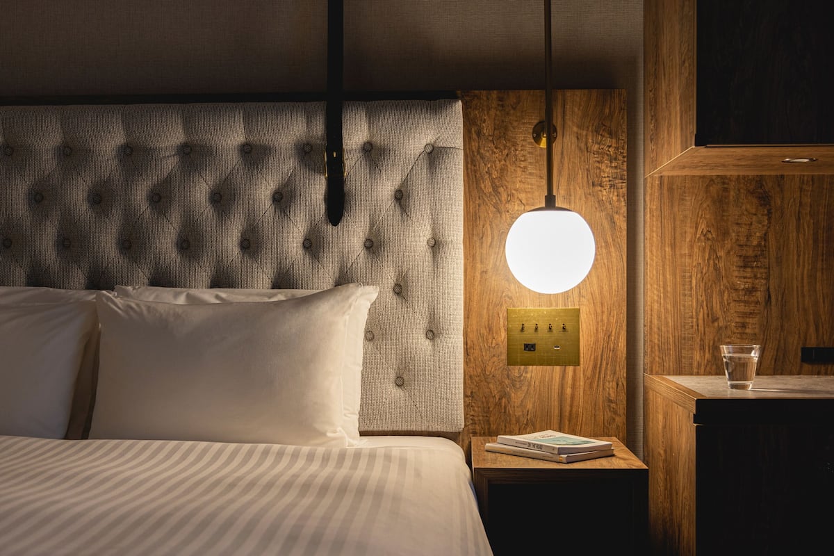 Un lit bien fait, avec du linge de lit blanc, une tête de lit à motifs texturés, une table de chevet avec un livre et un luminaire fixé au mur.