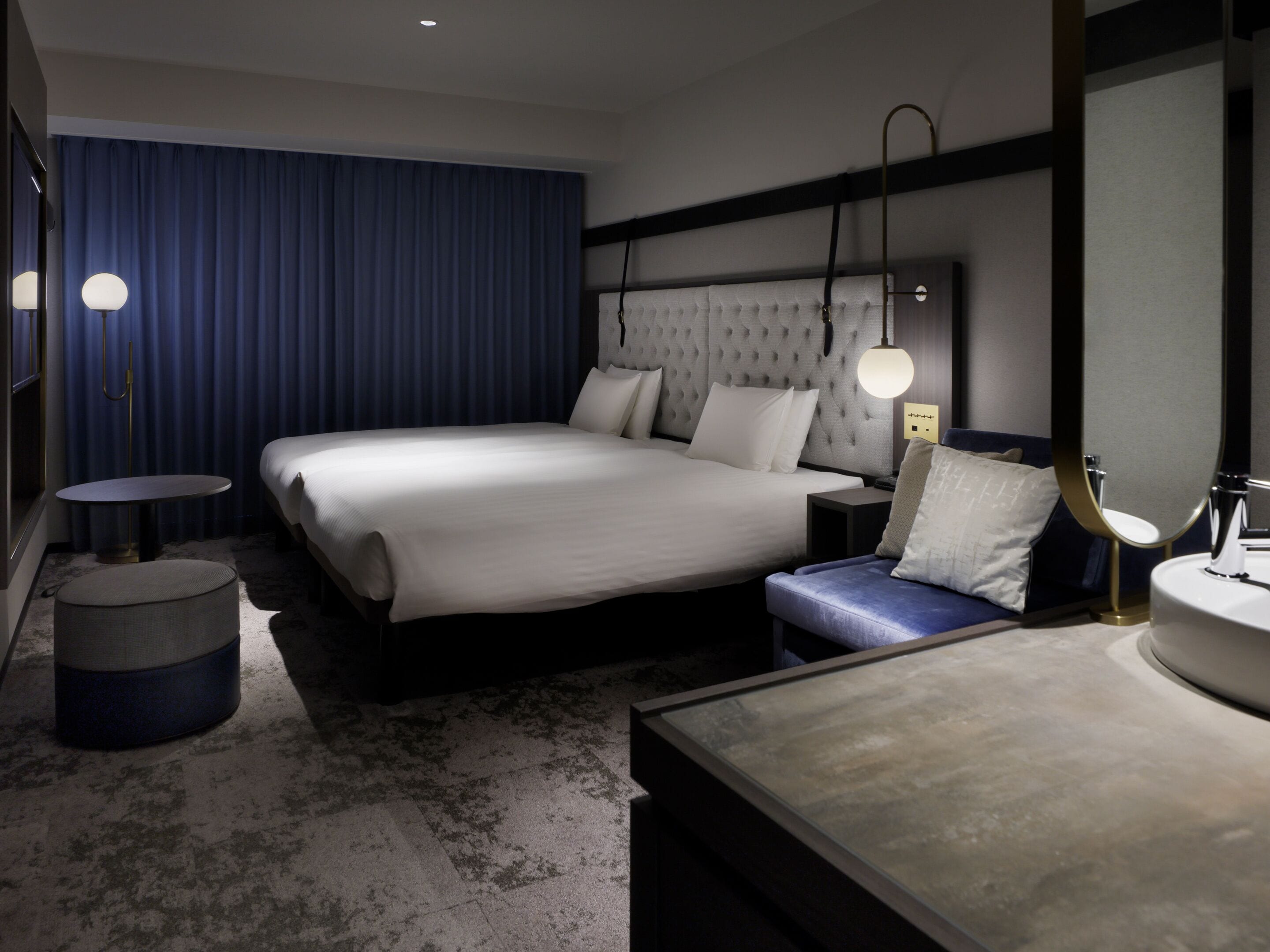 Hotelkamer met een groot bed, nachtkastjes, een rond poefje, een badkamer met een wastafel en spiegel, en een blauw gordijn.