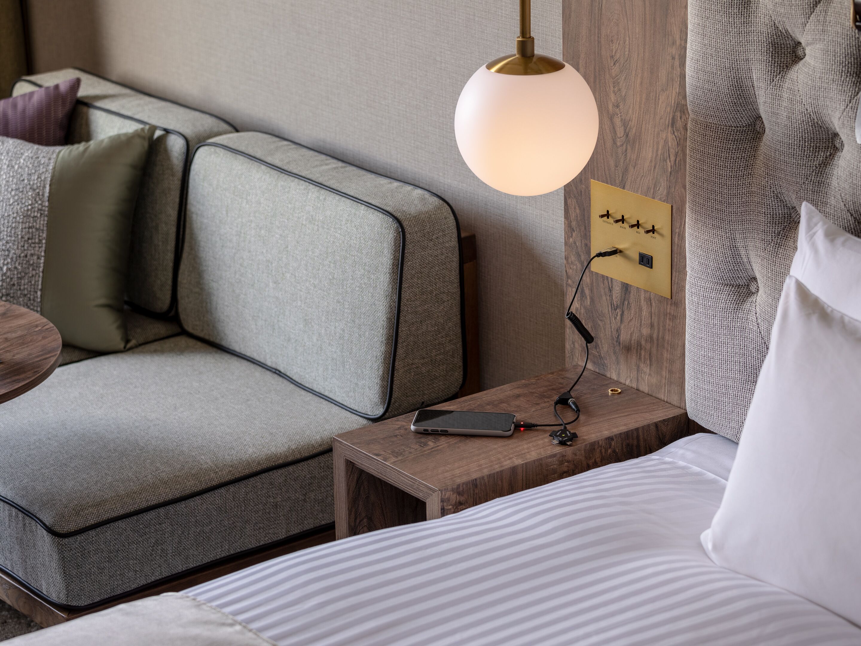 A modern hotel room with a grey sofa, a wooden nightstand with a lamp, and a bed with white bedding.