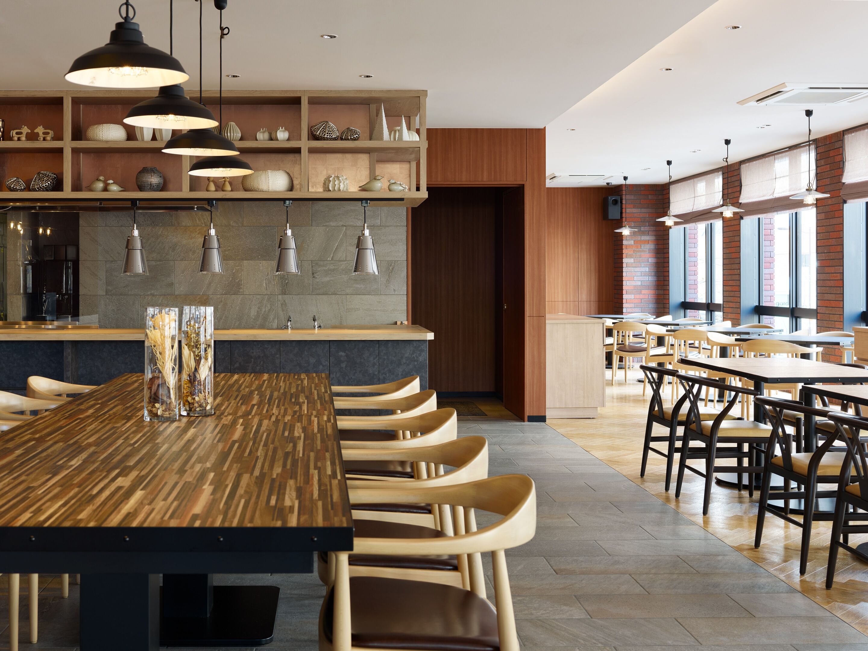 A modern restaurant with a long wooden table, multiple chairs, pendant lighting, and a brick wall.
