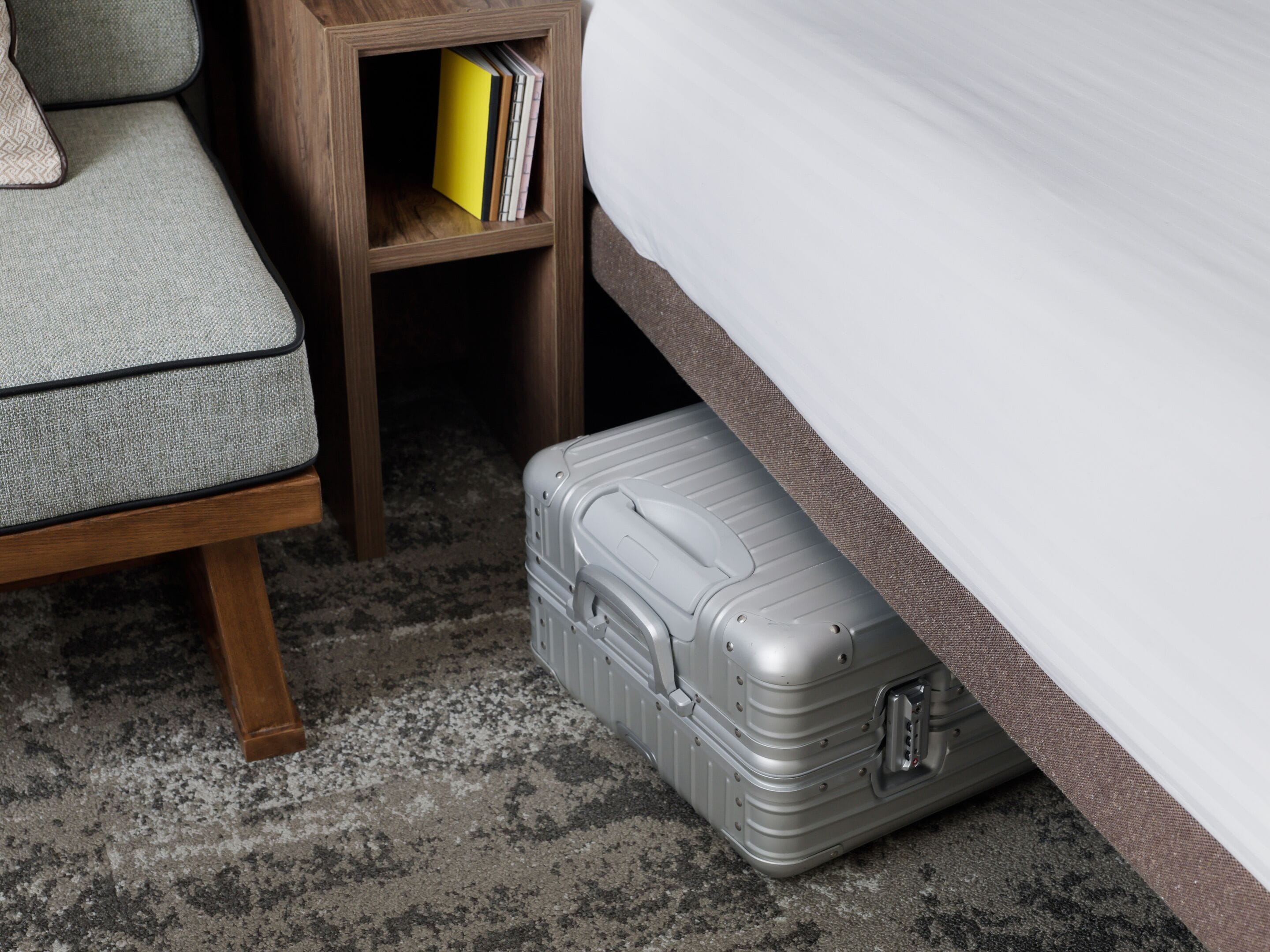 A bed, a wooden nightstand with books, and a grey suitcase on a carpeted floor.
