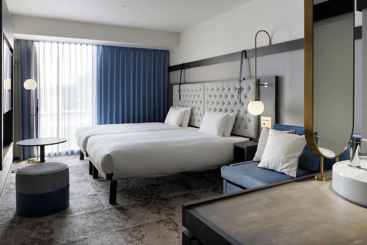 Une chambre d’hôtel moderne dotée d’un grand lit, de tables de chevet, d’un pouf rond, d’une salle de bain avec un lavabo et un miroir, et d’une grande fenêtre avec des rideaux.
