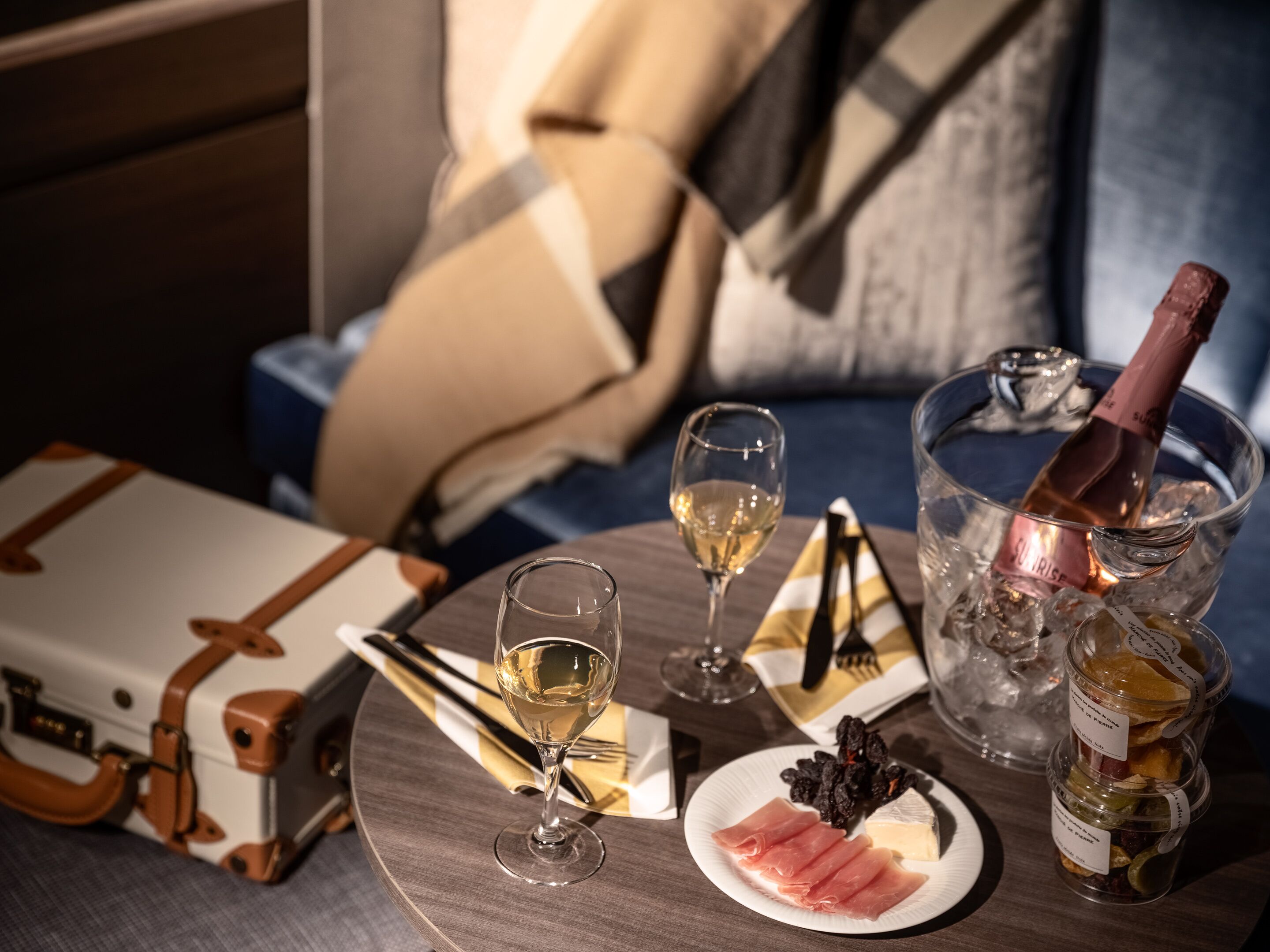 A dining table with a bottle of champagne, two glasses of wine, a plate of food, and a suitcase.