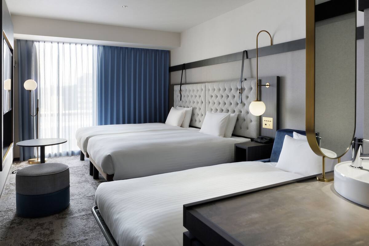 Une chambre d’hôtel avec deux lits, une table ronde, un pouf rond, une table de chevet avec une lampe, un grand miroir et une salle de bain avec un lavabo et un miroir.