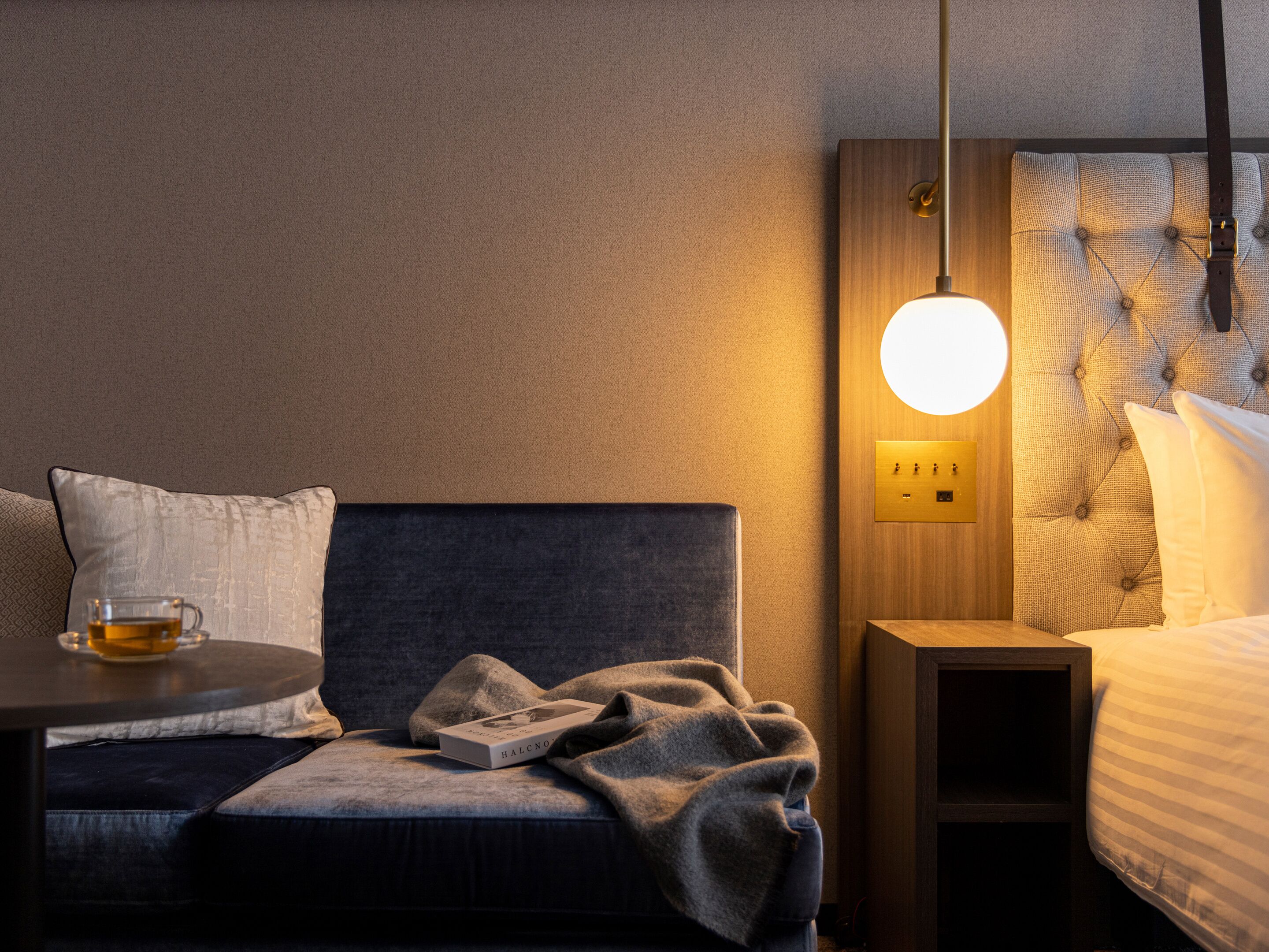 A hotel room with a bed, bedside table, and a reading area with a chair and a table.