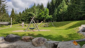 Un'area giochi con altalene, uno scivolo e una struttura con altalene, immersa nel verde e sullo sfondo alberi.
