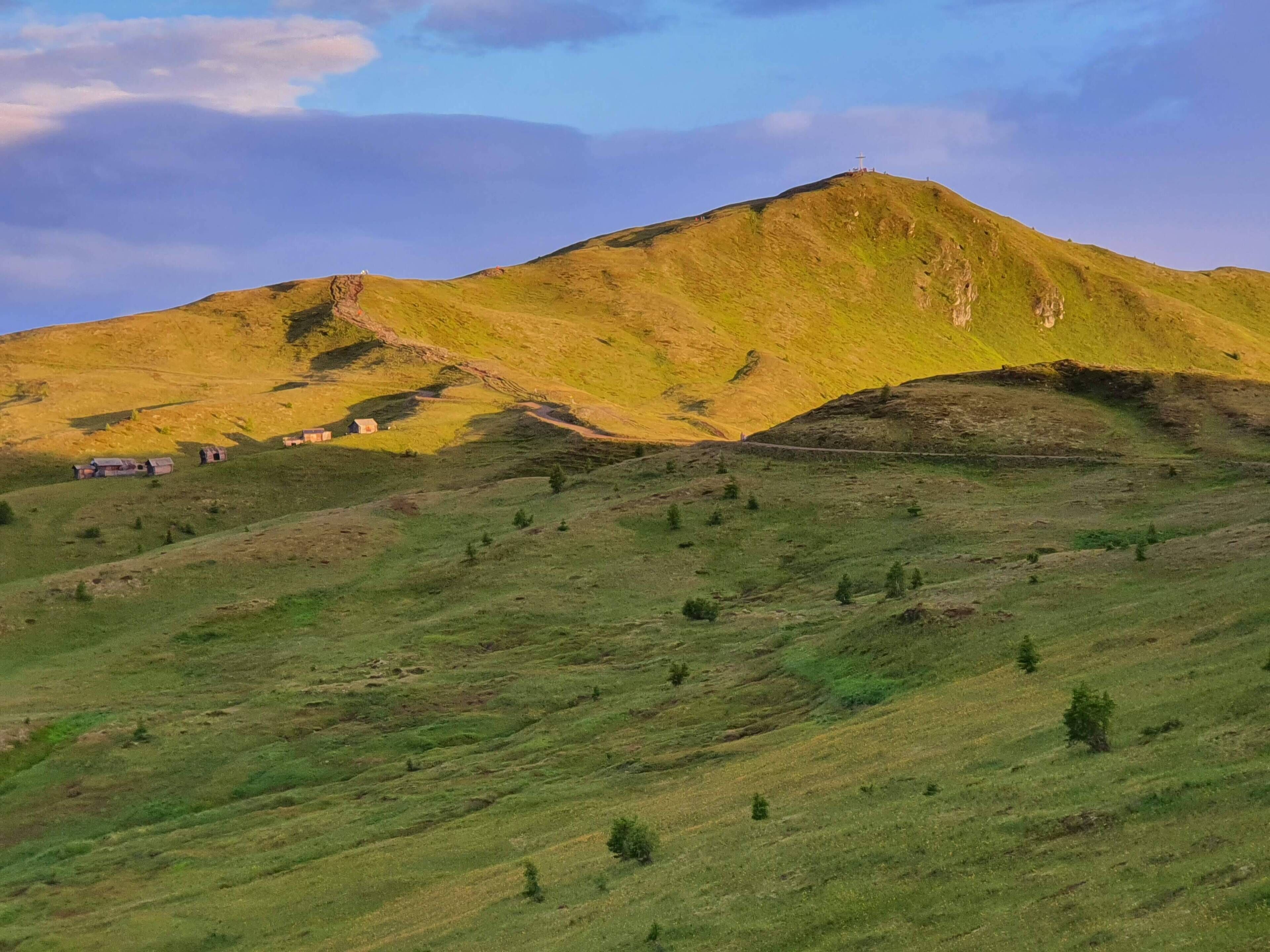 A mountainous landscape with a cross on top, a few buildings at the base, and a clear sky.