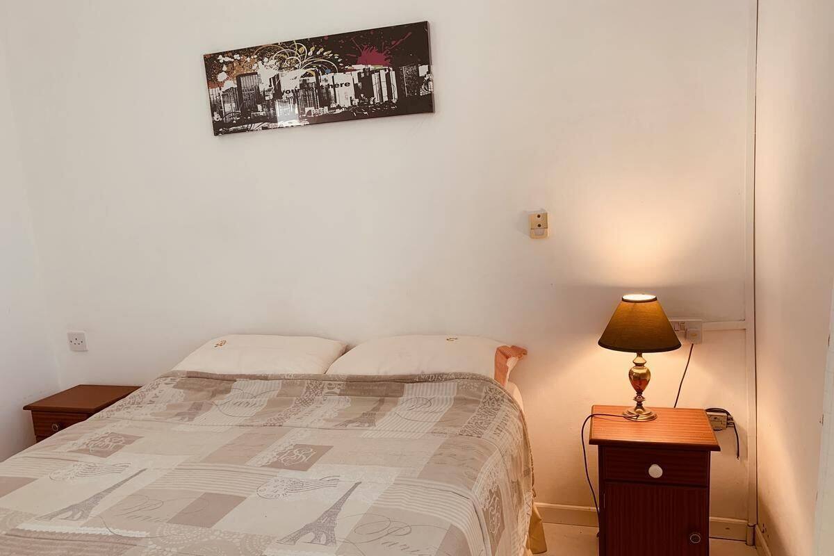 A bedroom with a bed, a nightstand with a lamp, a wall-mounted picture, and a small wooden table.