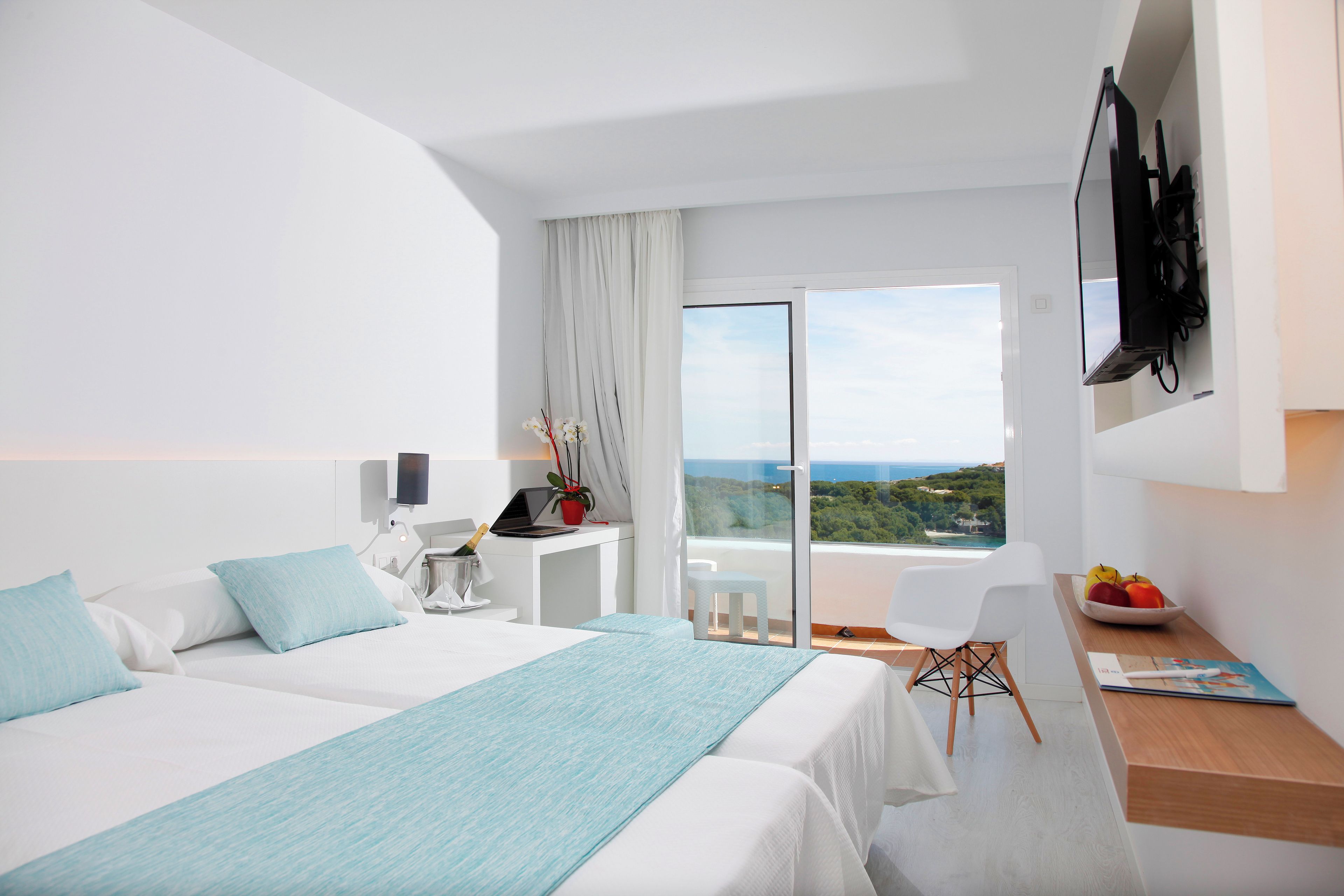 A modern hotel room with a bed, a desk, a chair, a TV, and a balcony with a view.