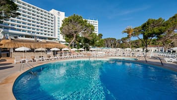 A large pool with lounge chairs and umbrellas, surrounded by a hotel building with multiple floors and a clear blue sky.