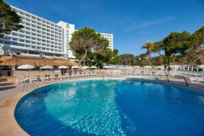 A large pool with lounge chairs and umbrellas, surrounded by a hotel building with multiple floors and a clear blue sky. - Aluasoul Hotel Carolina (Capdepera)