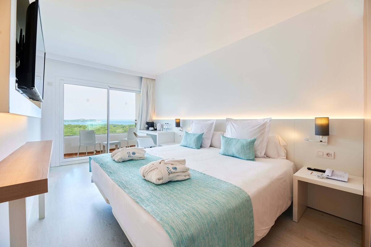 A hotel room with a large bed, a desk, a chair, a TV, and a balcony with a view.