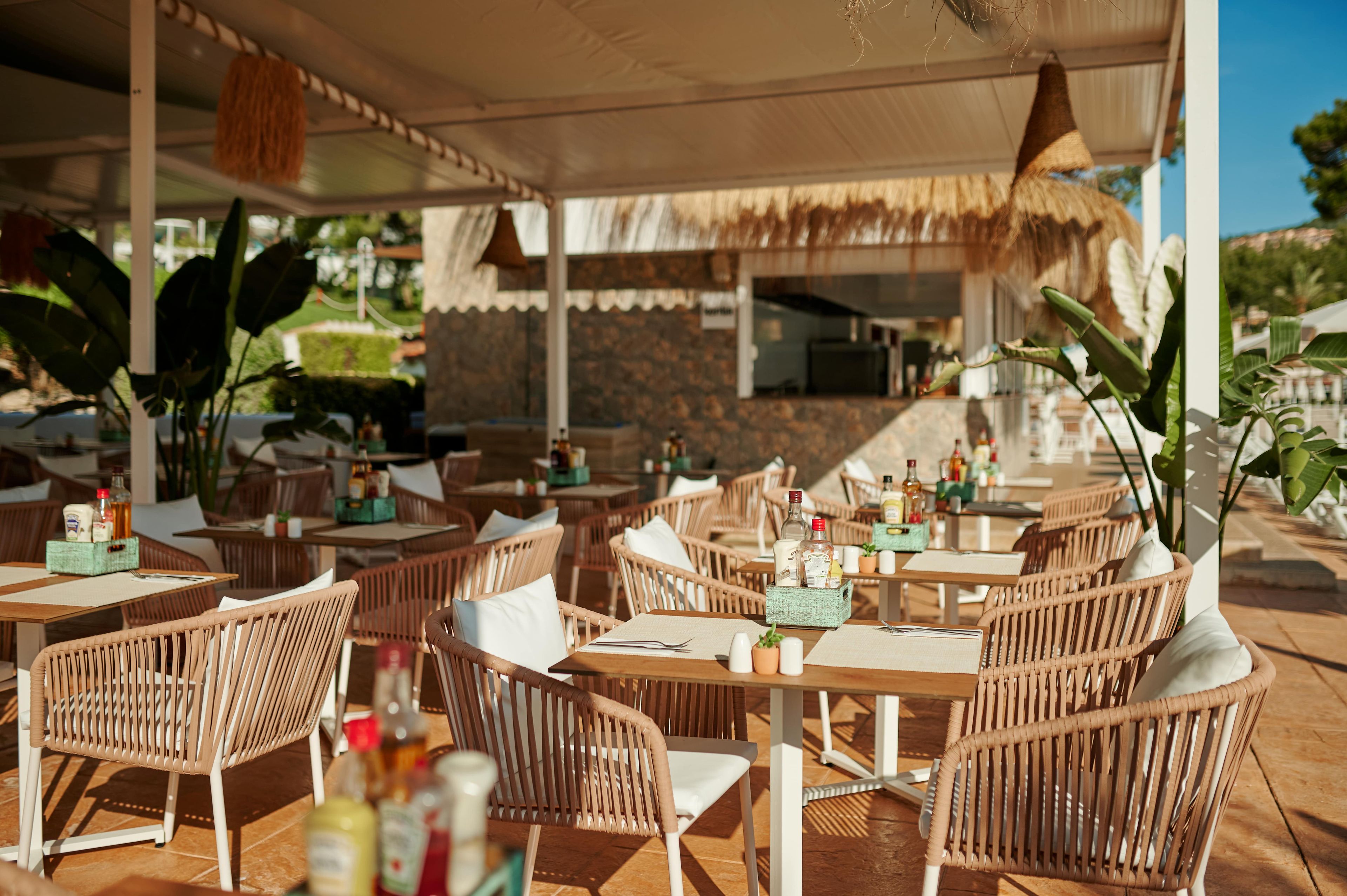 Een openluchtrestaurant met rieten meubels en een rieten dak. Tafels staan klaar met flessen en glazen.