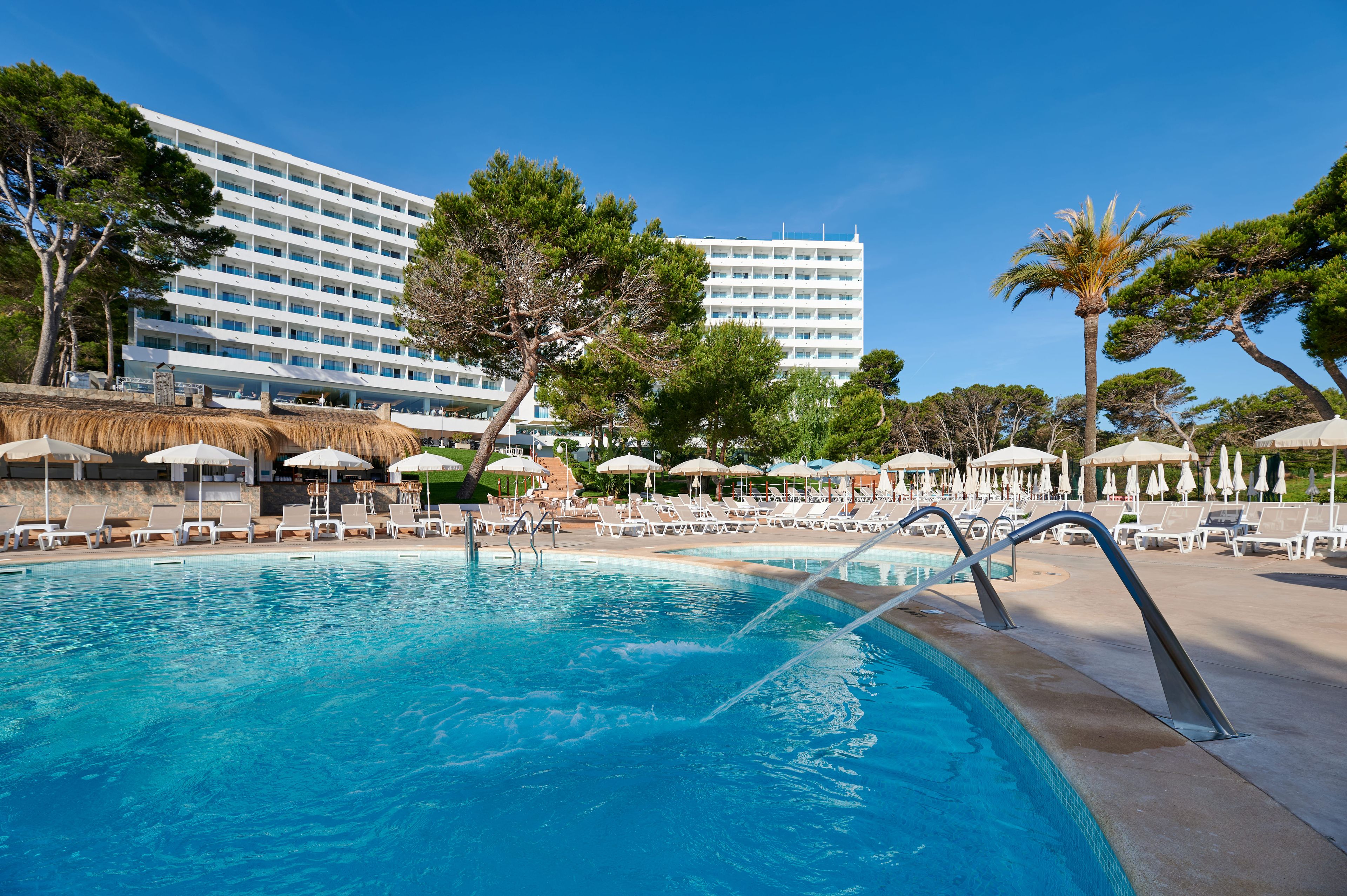 A swimming pool with lounge chairs and umbrellas, surrounded by trees and a hotel building.